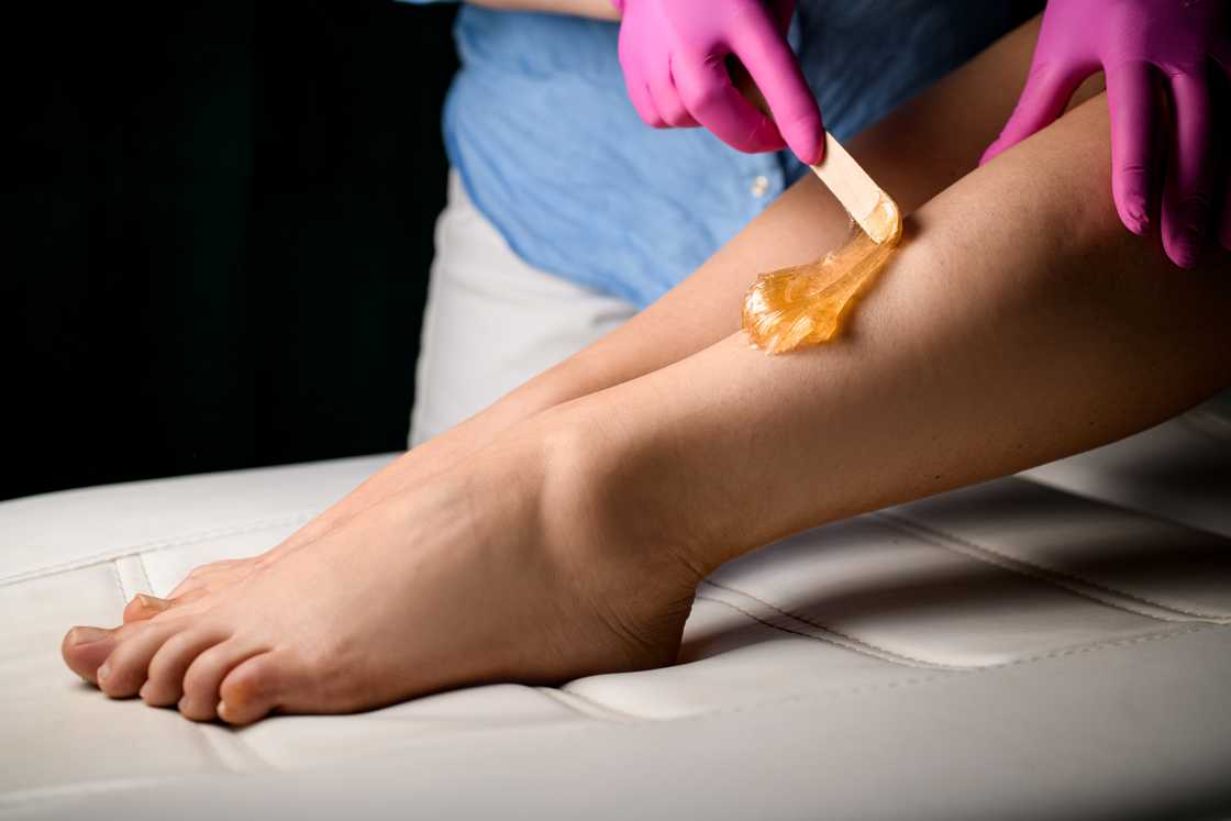 A sugaring specialist dressed in a blue shirt and pink rubber gloves applies sugar on the legs of a sitting woman