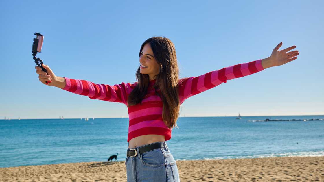 A happy young woman records herself with a gimbal at the sunny seaside, arms wide open