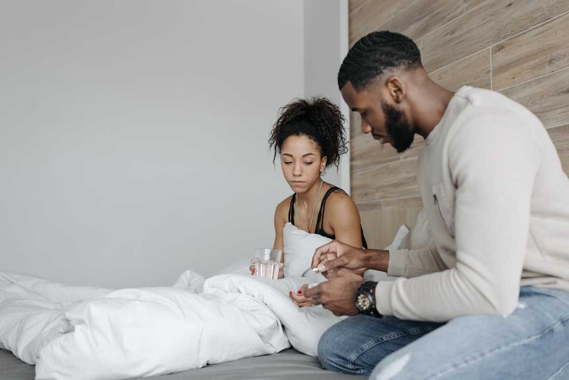 A man helps his unwell partner by preparing medicine while she rests in bed.