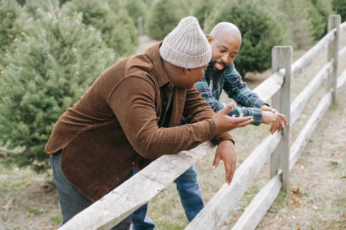 Two men lean on a wooden fence outdoors, talking and sharing a relaxed conversation.