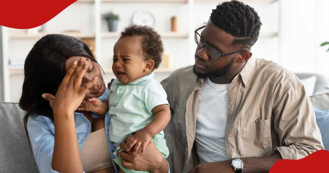 Photo of black mum and partner sitting with a small crying black kid on couch at home.