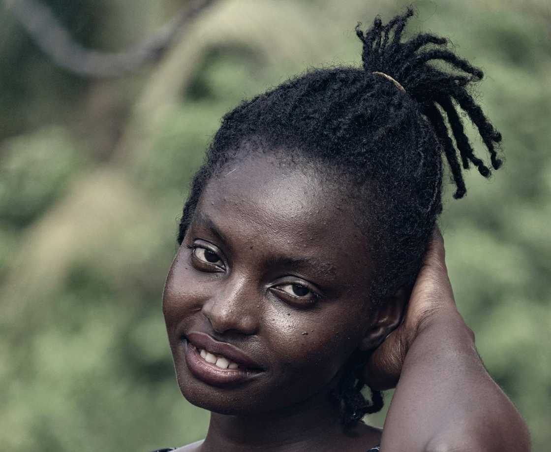 A smiling woman outdoors with her hair tied back, touching her head.