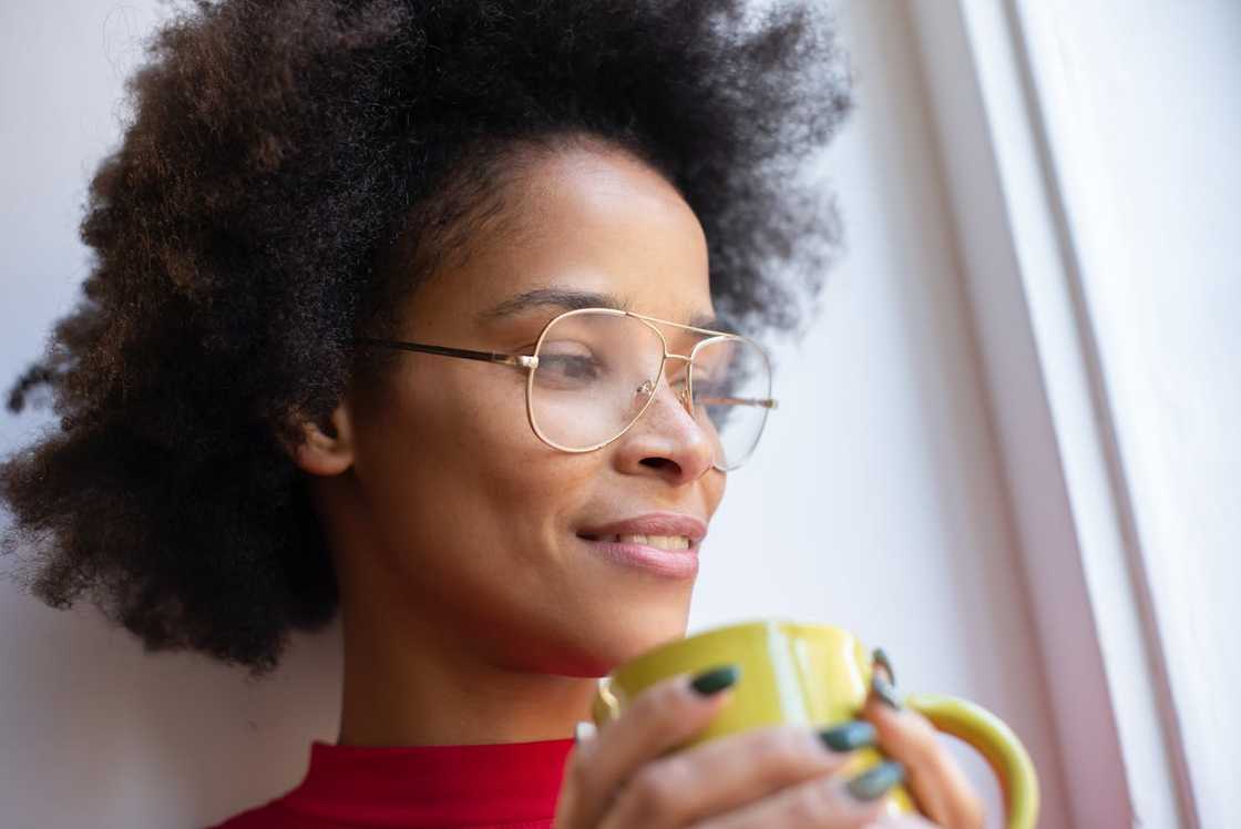 A woman with glasses holding a mug and looking out a window.