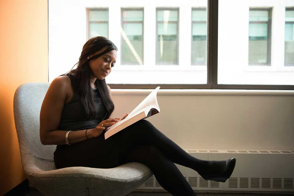 A woman in an all-black outfit reading a book by the window