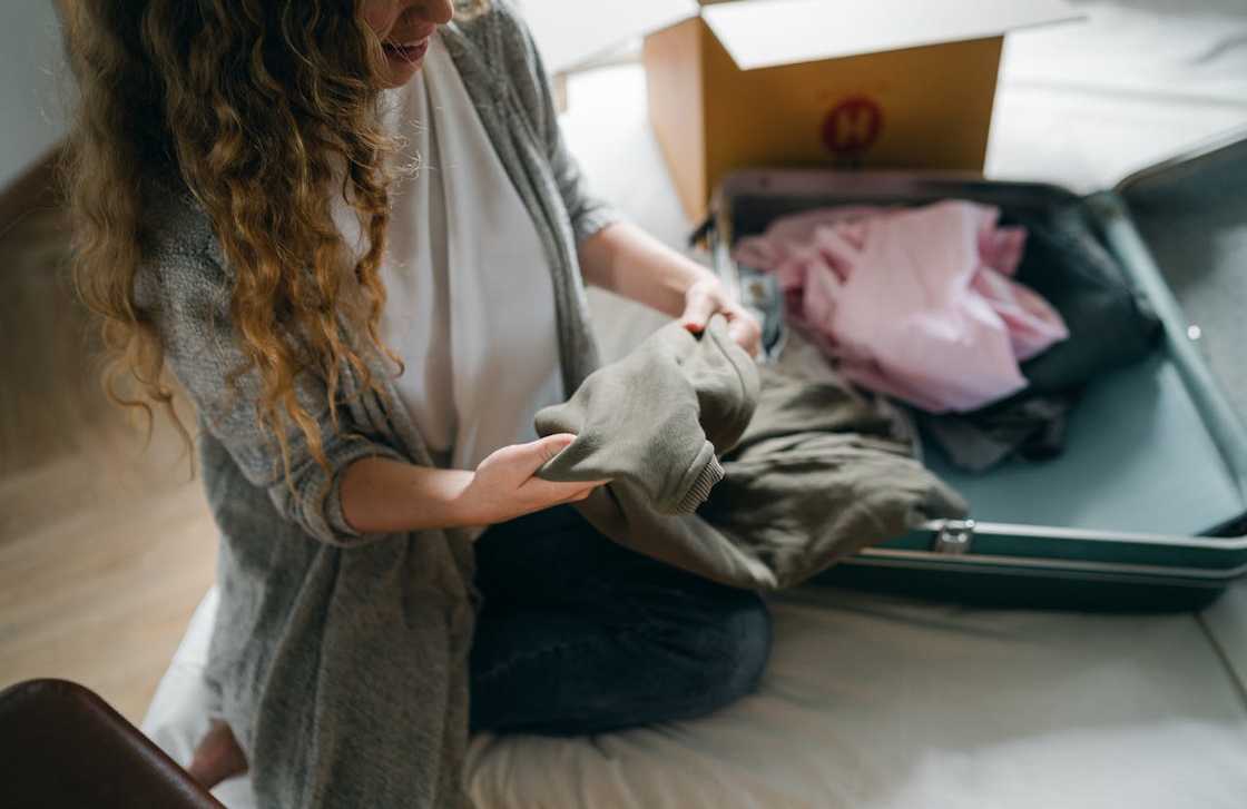 A woman kneels on the floor packing clothes into an open suitcase.