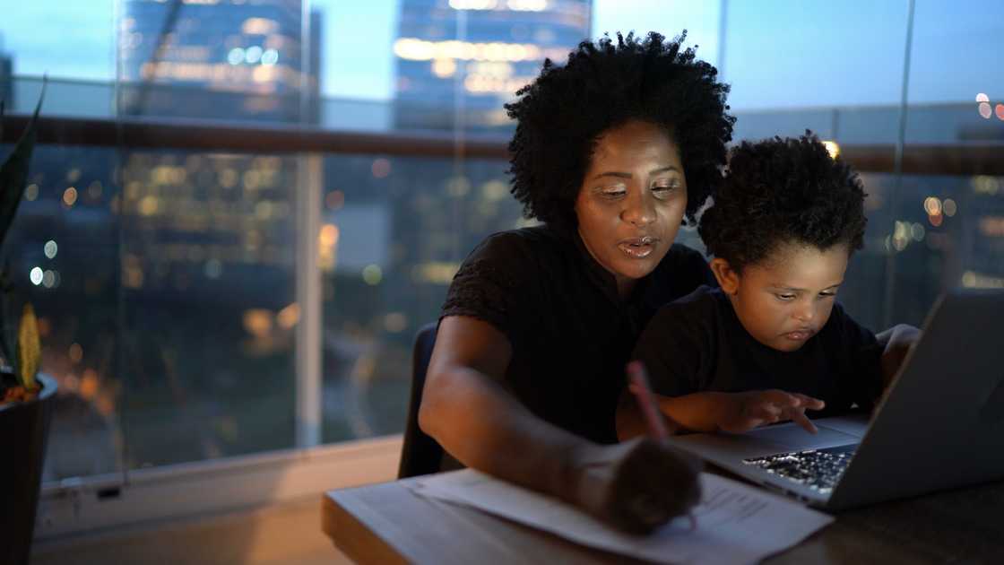 Adult and child sit at a table with a laptop, working together as city lights glow outside. Adult and child sit at a table with a laptop, working together as city lights glow outside.