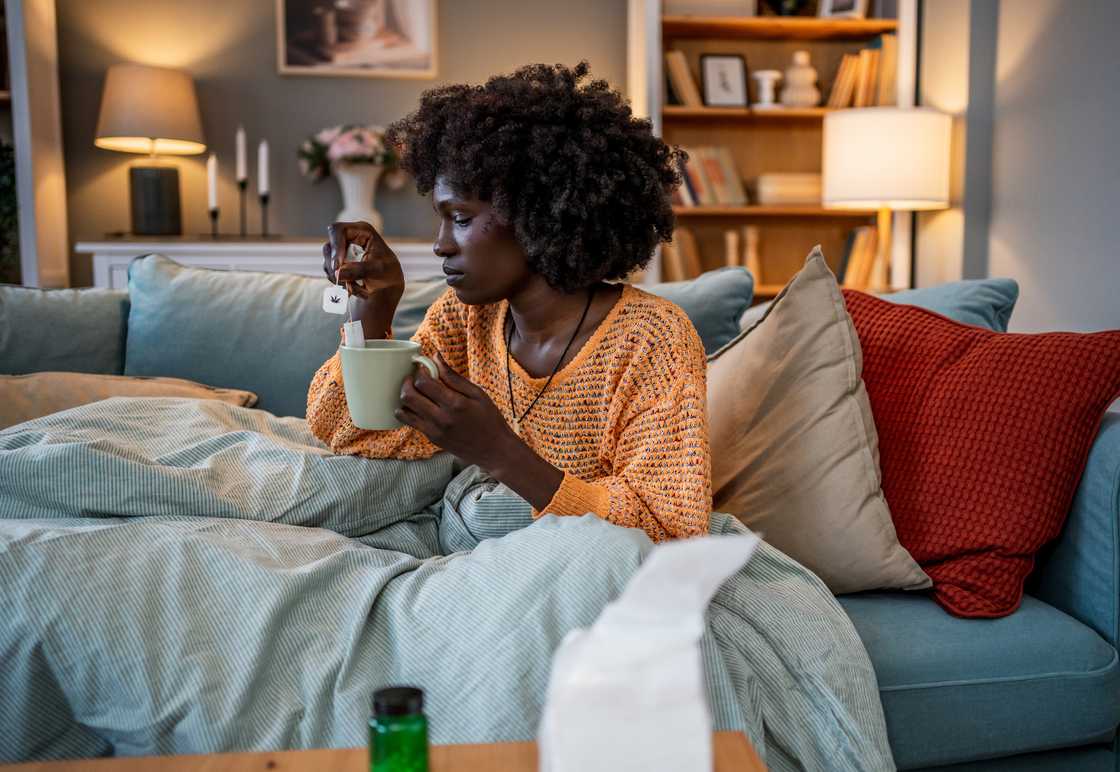 Person on couch with blanket and tea, medicine and tissues nearby.