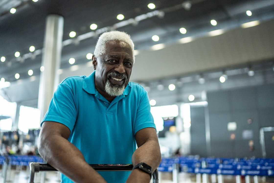 Older person in a bright blue shirt smiles while leaning on a luggage cart in a modern transportation terminal. Older person in a bright blue shirt smiles while leaning on a luggage cart in a modern transportation terminal.