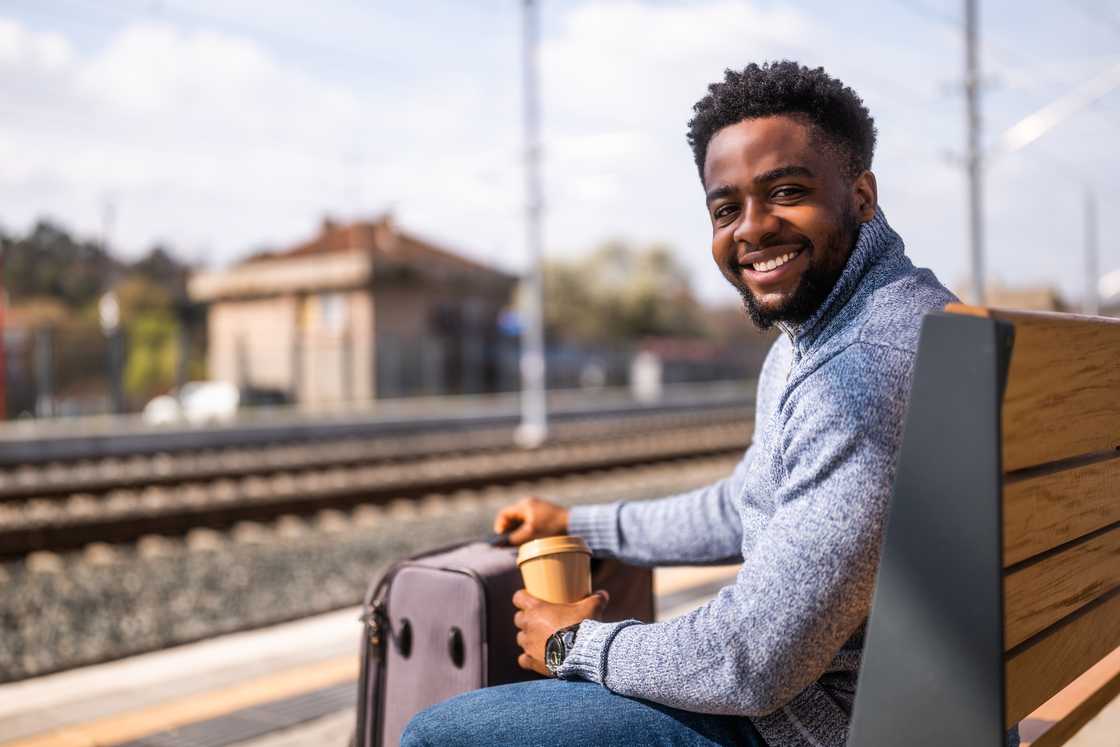 Person sits on a wooden bench at a train station, smiling and holding a coffee cup with a suitcase beside them. Person sits on a wooden bench at a train station, smiling and holding a coffee cup with a suitcase beside them.