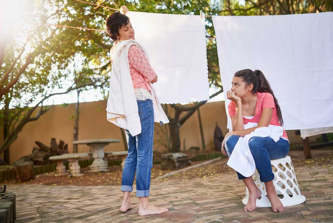 Two barefoot individuals in jeans and pink tops interact near a backyard clothesline with white sheets.
