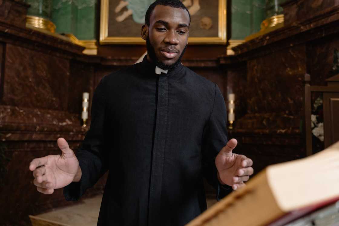 A man in clerical robe standing indoors with hands raised in a prayerful gesture.