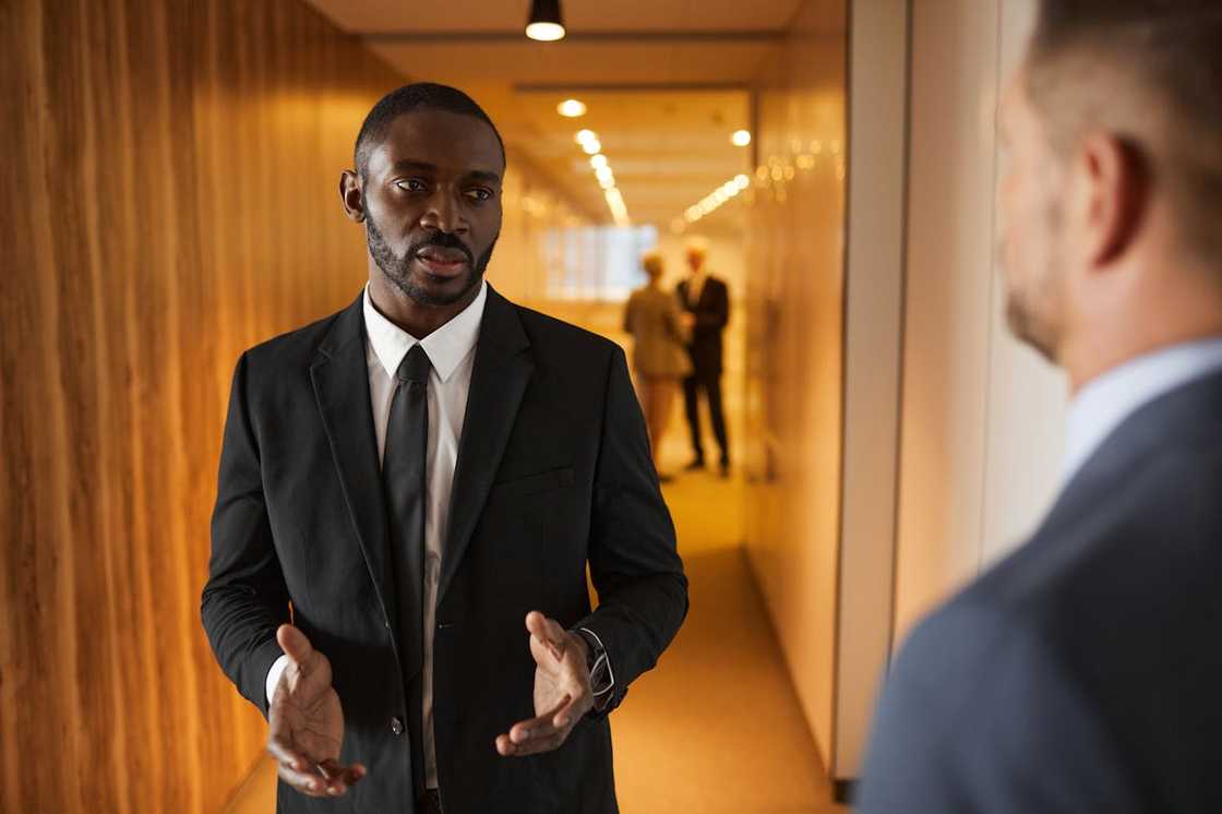 A man in a suit speaks to another man in a hallway.