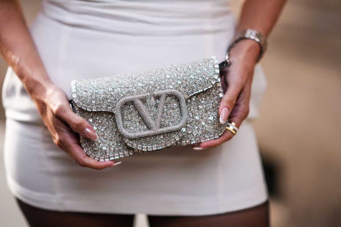 A woman in a white dress elegantly holds a silver Valentino clutch bag adorned with sparkling rhinestones. A woman in a white dress elegantly holds a silver Valentino clutch bag adorned with sparkling rhinestones.