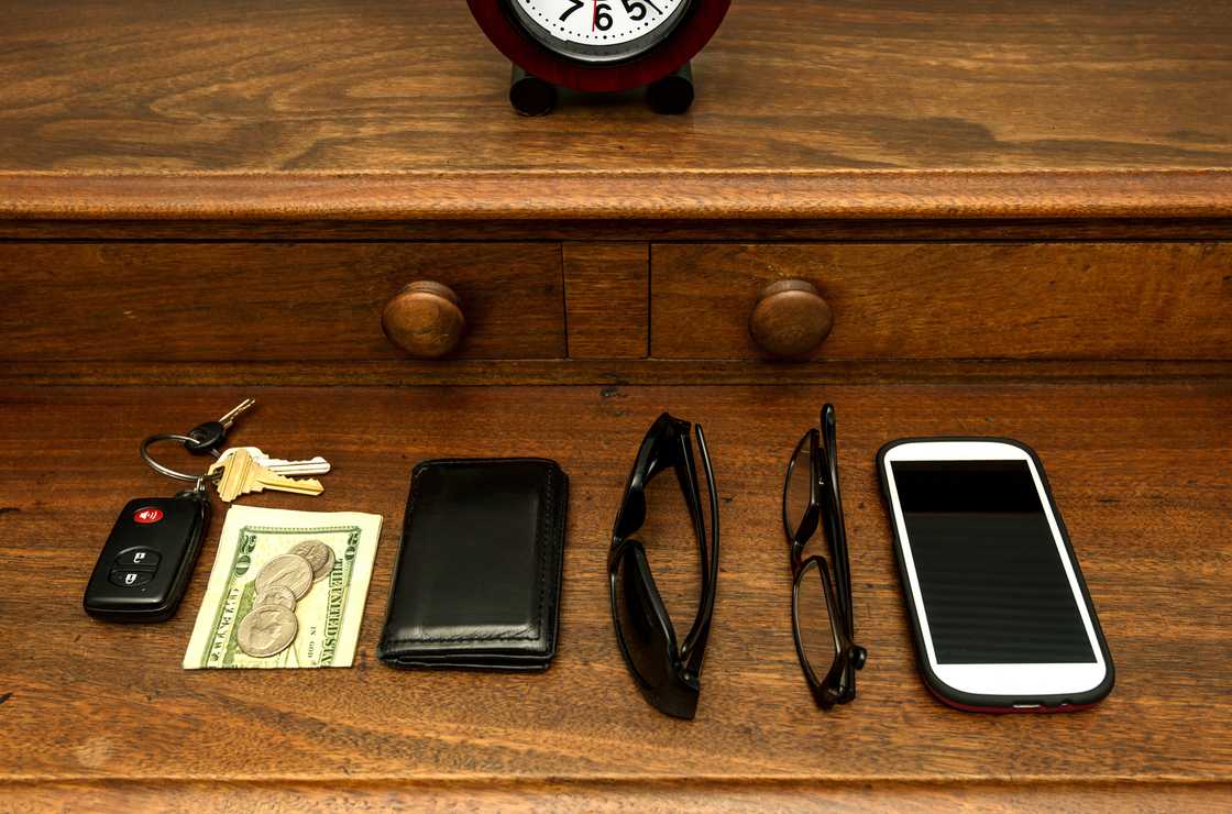 A phone, wallet, shades, car keys, and money on top of a dresser