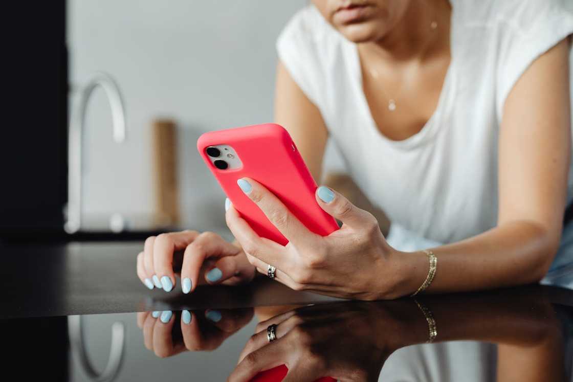 A woman wearing glasses looks down at her phone while standing against a light wooden wall.
