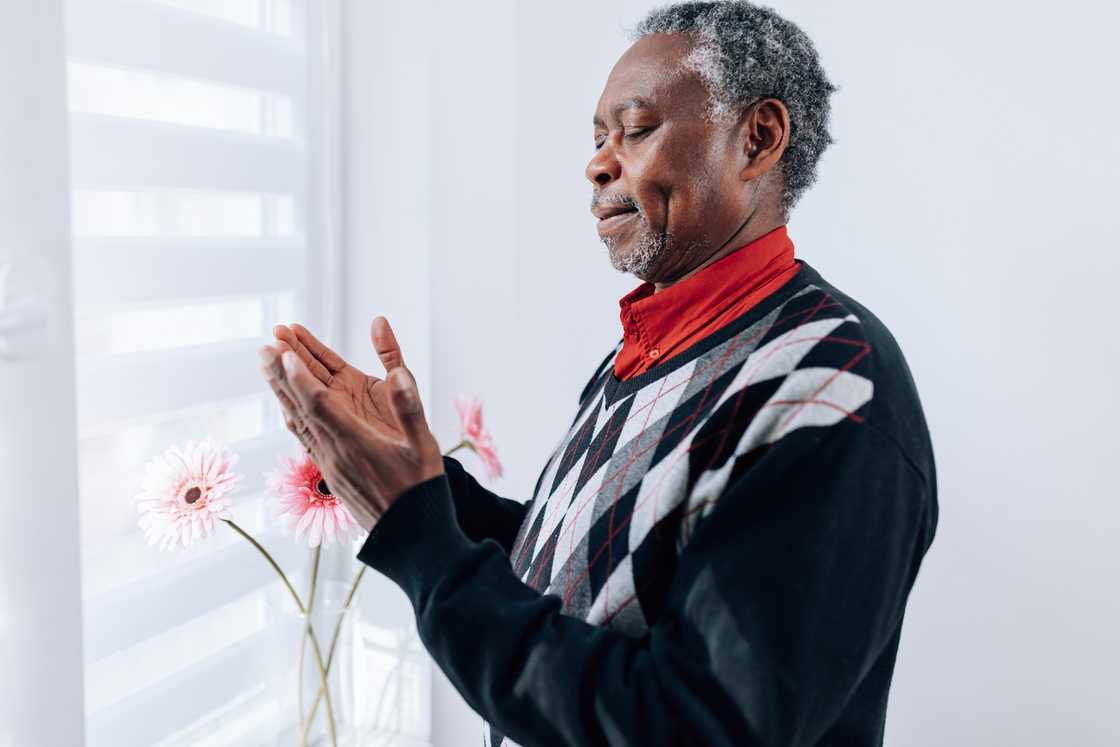 Older person stands indoors near a window with hands gently clasped.
