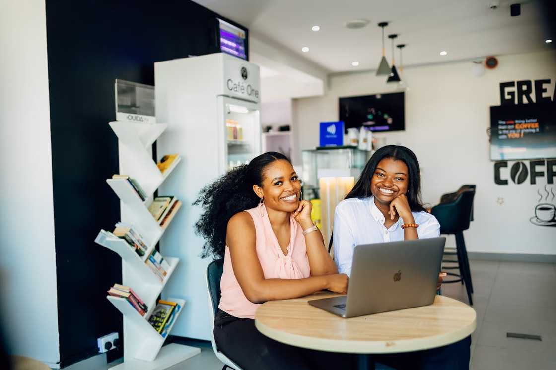 Two women smiling while working together on a laptop at a café table. Two women smiling while working together on a laptop at a café table.