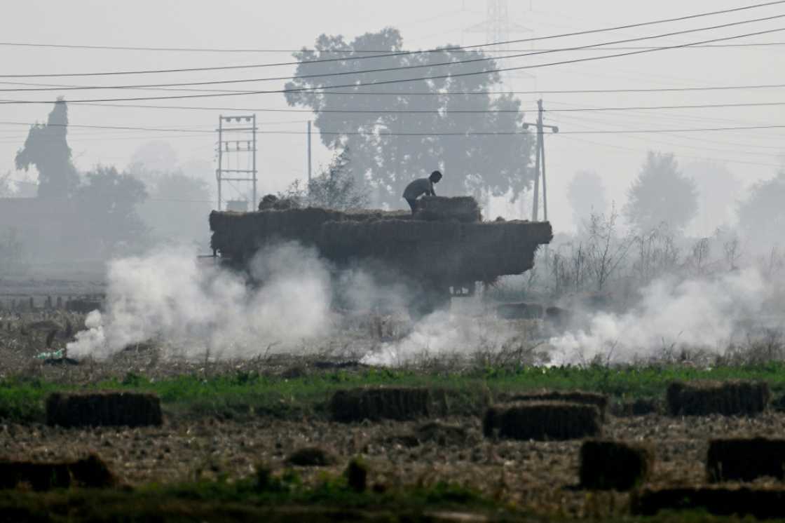 A worker loads a tractor with paddy straw bales as smoke rises from smoldering leftover stubble A worker loads a tractor with paddy straw bales as smoke rises from smoldering leftover stubble