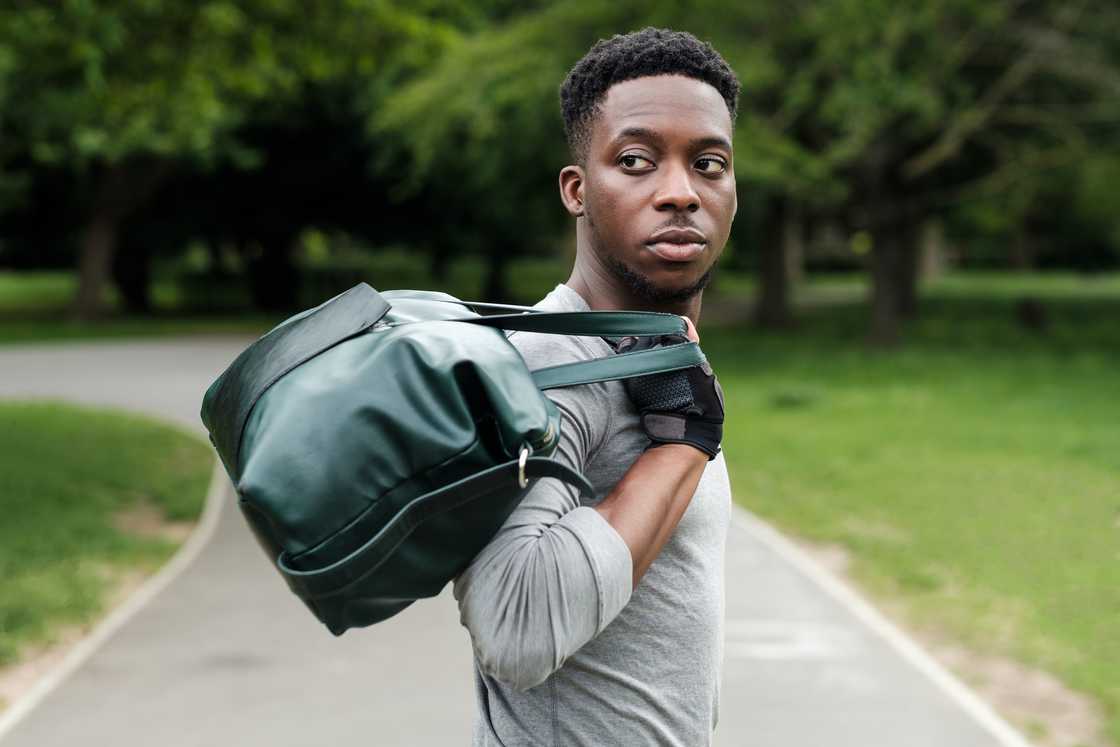 A young man walks on a paved path carrying a duffel bag in a park setting.