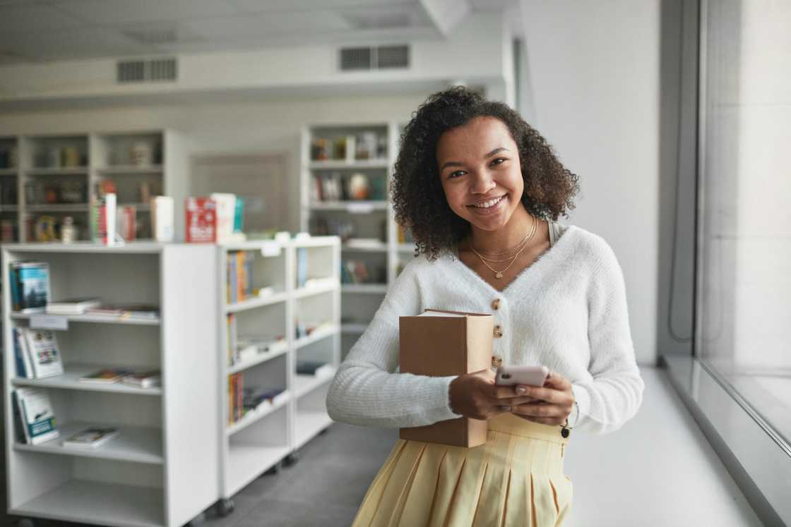 A woman in a white sweater and brown skirt holding a book and a smartphone A woman in a white sweater and brown skirt holding a book and a smartphone