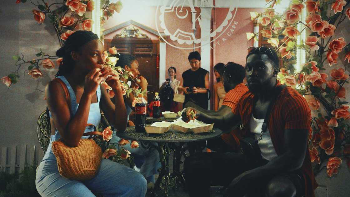 People sit at a small café table, eating and chatting among floral decorations.