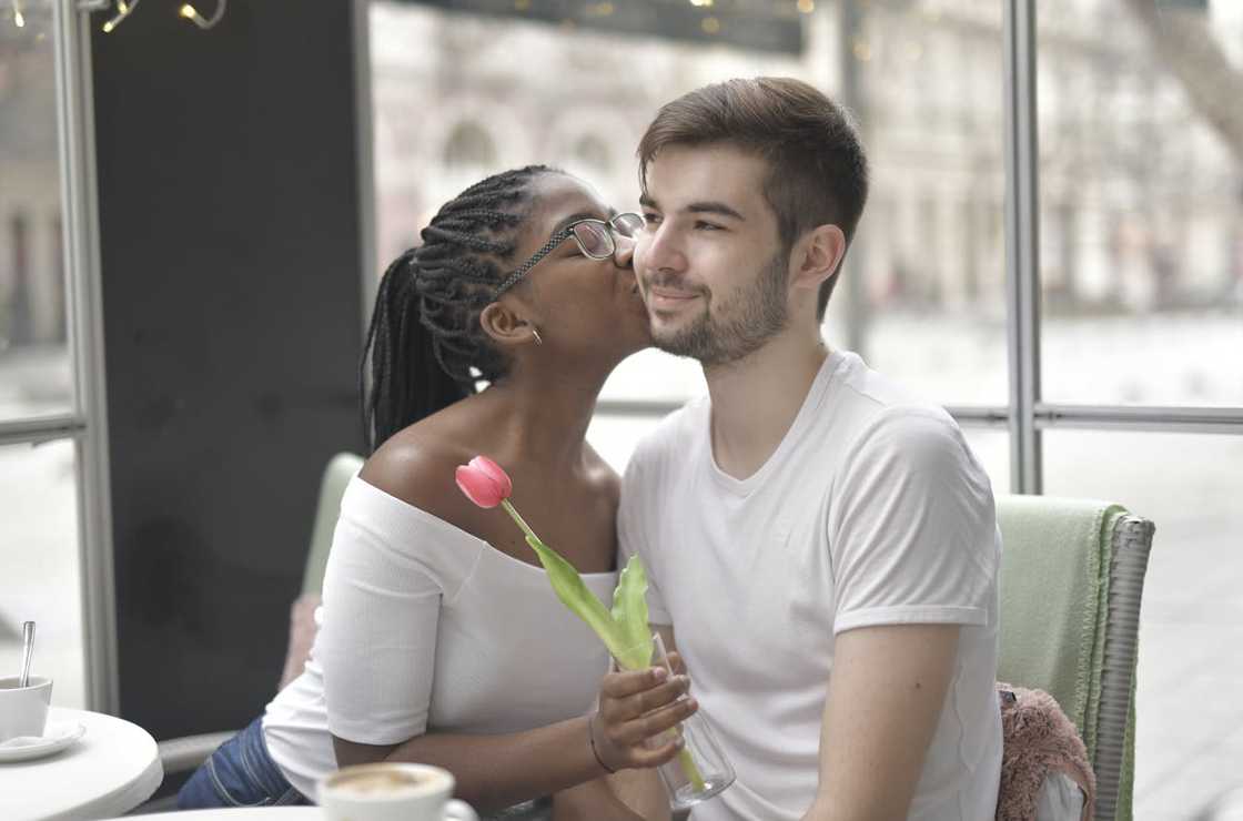 A woman kisses a man on the cheek while holding a single flower in a café.
