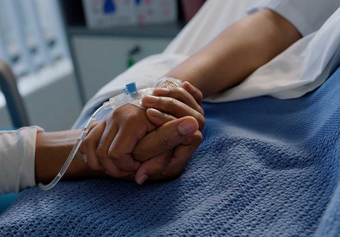Two hands clasp together in a hospital bed—one with an IV line, the other offering comfort. Two hands clasp together in a hospital bed—one with an IV line, the other offering comfort.